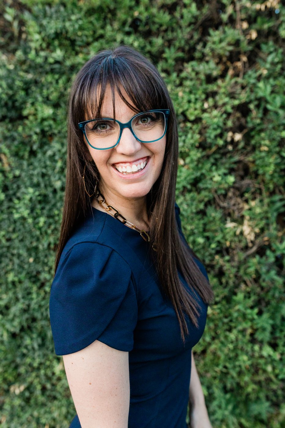 Sara Jane smiling, wearing a navy top, standing in front of green foliage